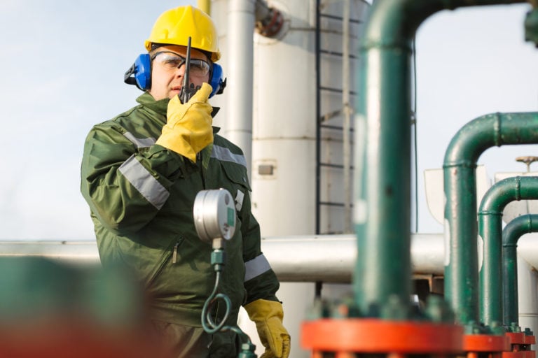 A gas production operator wears safety equipment and uses a 2-way radio while standing outside an oil plant.