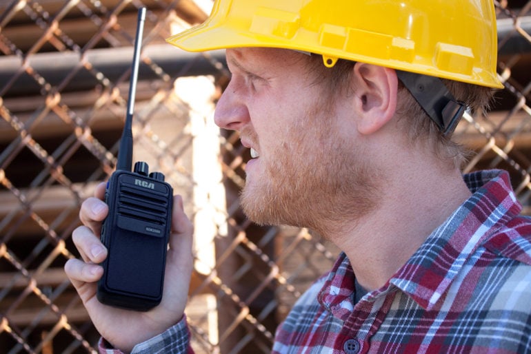 A construction worker in a hardhat uses an RCA RDR2500 two-way radio while standing next to a chain-link fence.